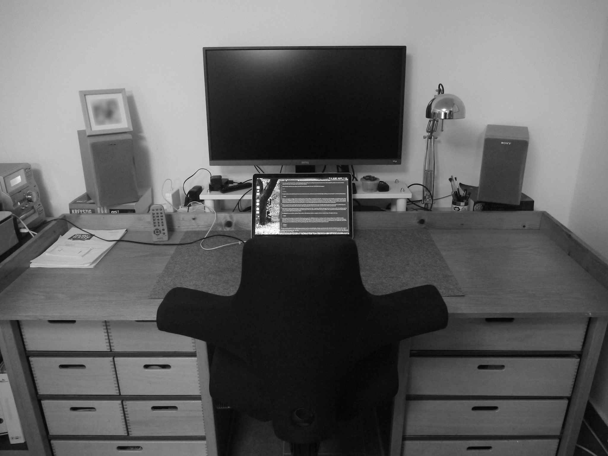Black and white photo of a simple wooden desk on two sets of draws. In the middle sits a laptop, and behind it a large black screen. Left and right are stereo speakers.