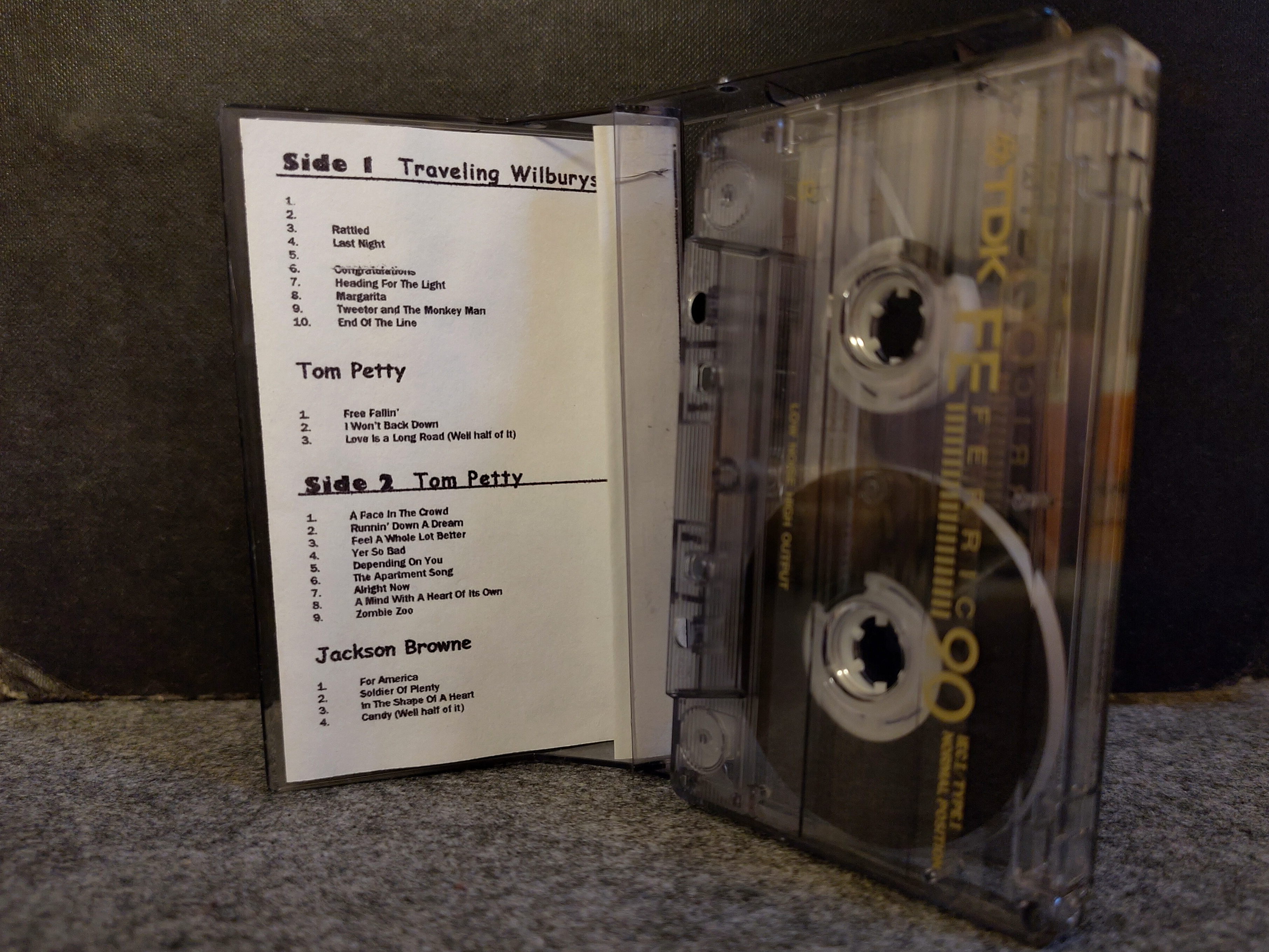Photo of an audio cassette, standing next to its open case. Inside there is a home printed paper insert listing the songs on the cassette.